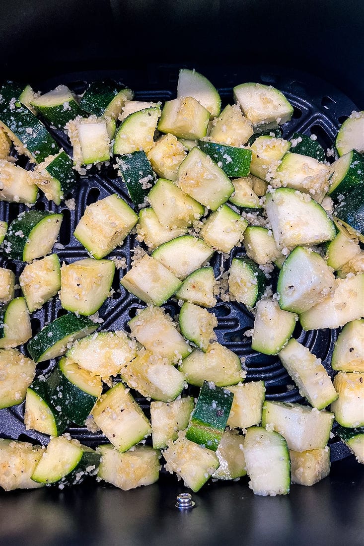 Cubes of raw zucchini, seasoned and covered in panko breadcrumbs and parmesan cheese, in an air fryer basket.