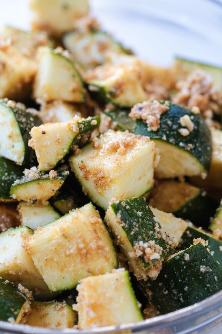 Close up of raw zucchini in a mixing bowl, coated in seasonings, including parmesan cheese and panko breadcrumbs