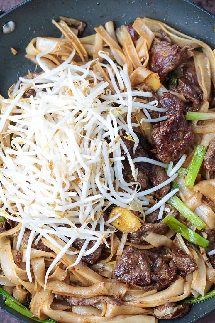 A pile of bean sprouts resting atop beef chow fun in a pan