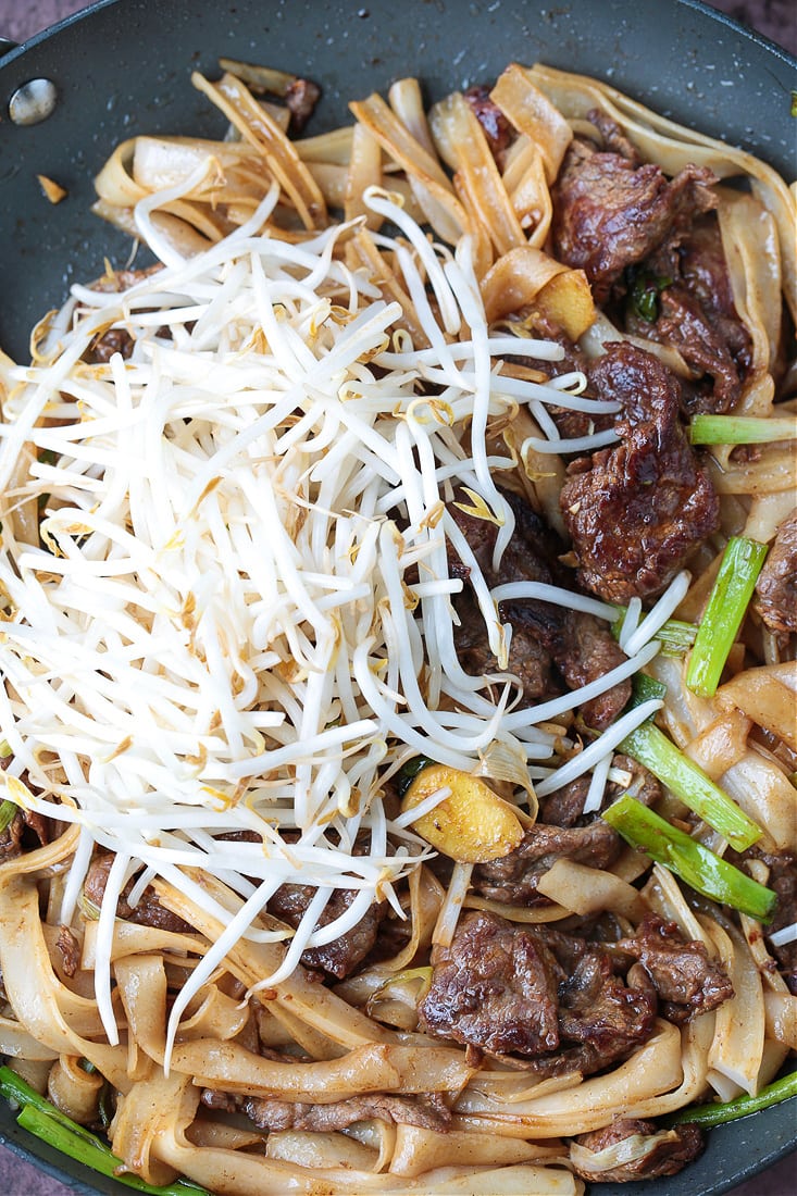 This beef chow fun recipe is a classic Chinese takeout dish, with marinated flank steak, green onions, rice noodles, and bean sprouts. A pile of bean sprouts resting atop beef chow fun in a pan