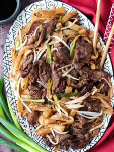 An oval serving tray filled with beef chow fun, with chopsticks in the platter and scallions next to it