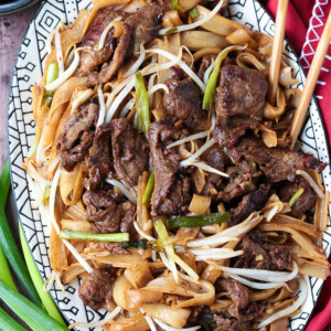 An oval serving tray filled with beef chow fun, with chopsticks in the platter and scallions next to it