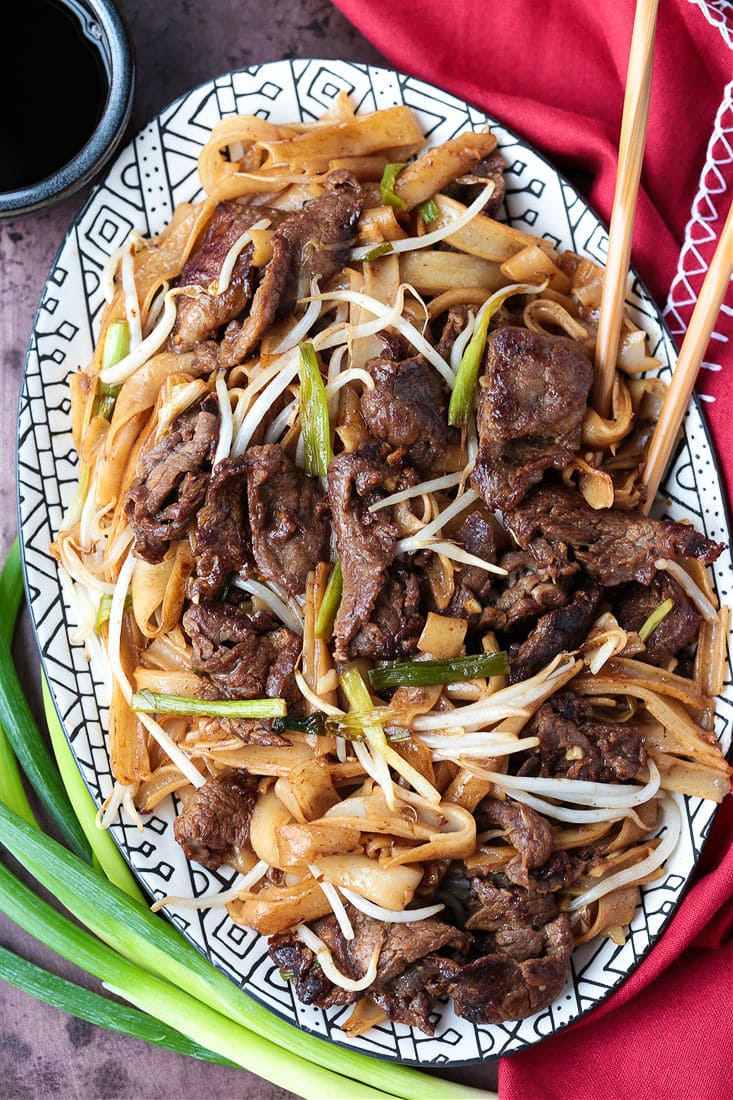 This beef chow fun recipe is a classic Chinese takeout dish, with marinated flank steak, green onions, rice noodles, and bean sprouts. An oval serving tray filled with beef chow fun, with chopsticks in the platter and scallions next to it