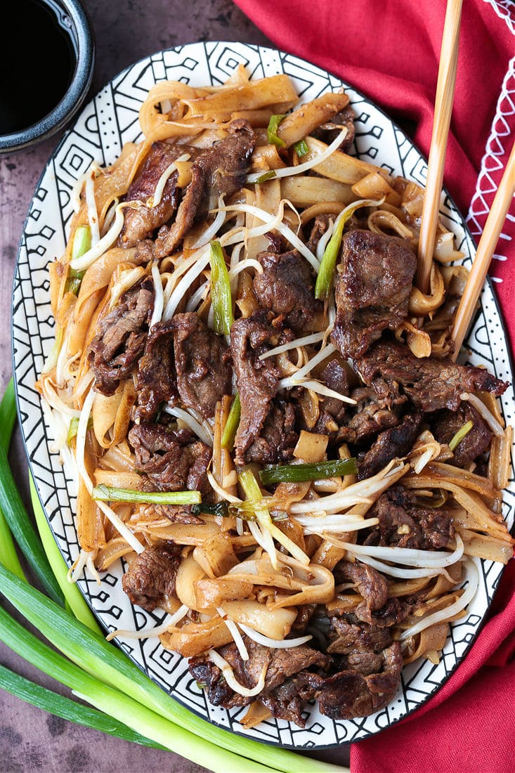 An oval serving tray filled with beef chow fun, with chopsticks in the platter and scallions next to it