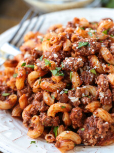 Close up of beefaroni on a plate, garnished with parsley and parmesan, with a fork in the background