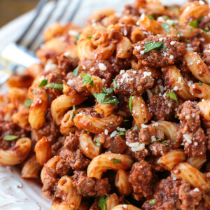 Close up of beefaroni on a plate, garnished with parsley and parmesan, with a fork in the background