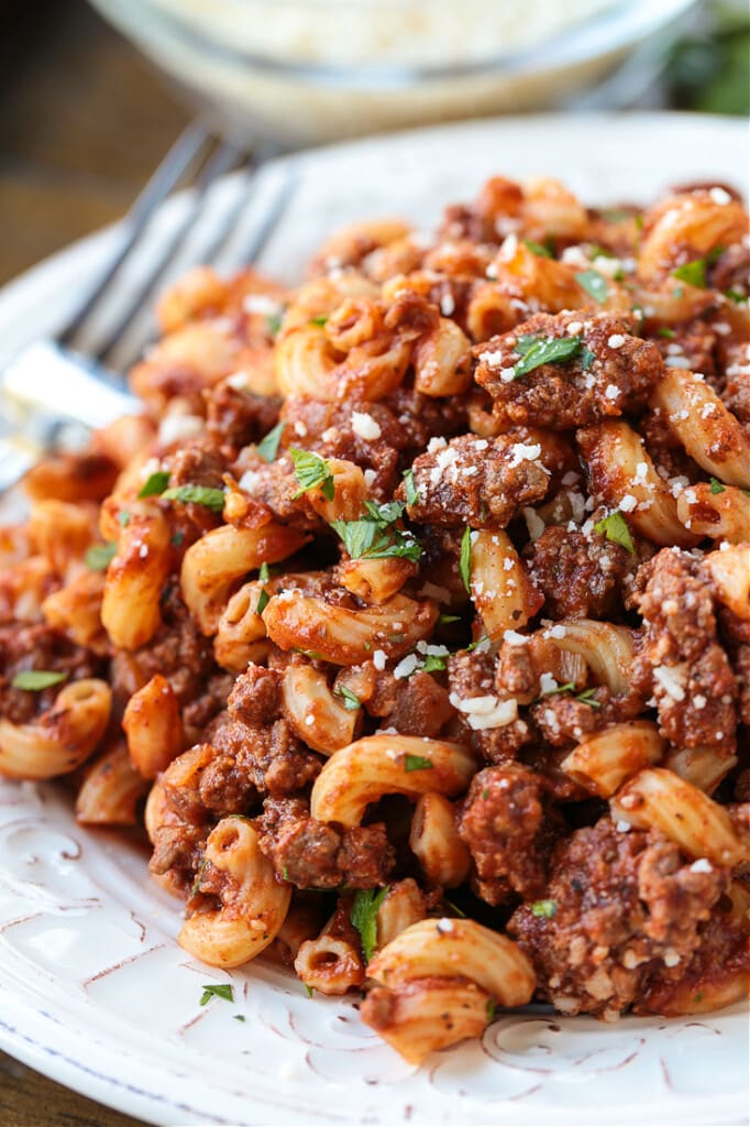 Close up of beefaroni on a plate, garnished with parsley and parmesan, with a fork in the background
