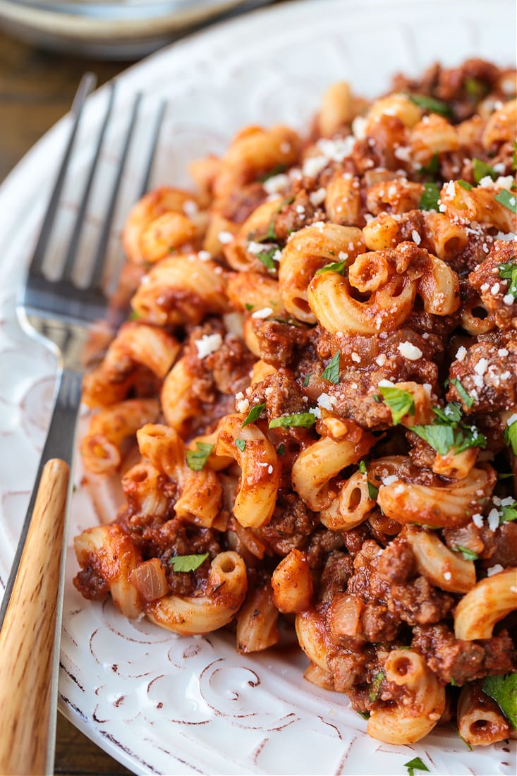 Close up of beefaroni on a plate, with parmesan and parsley on top, and a fork on the side
