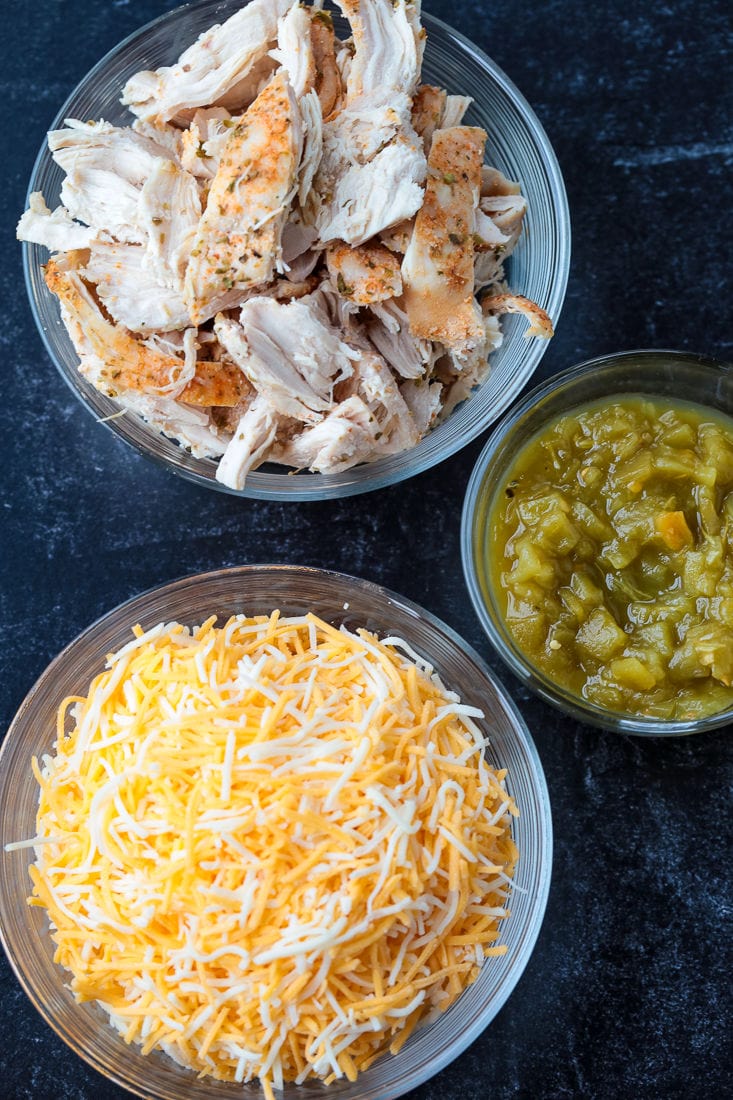 A bowl of cooked chicken next to a bowl of shredded cheese and a bowl of hatch green chilis 