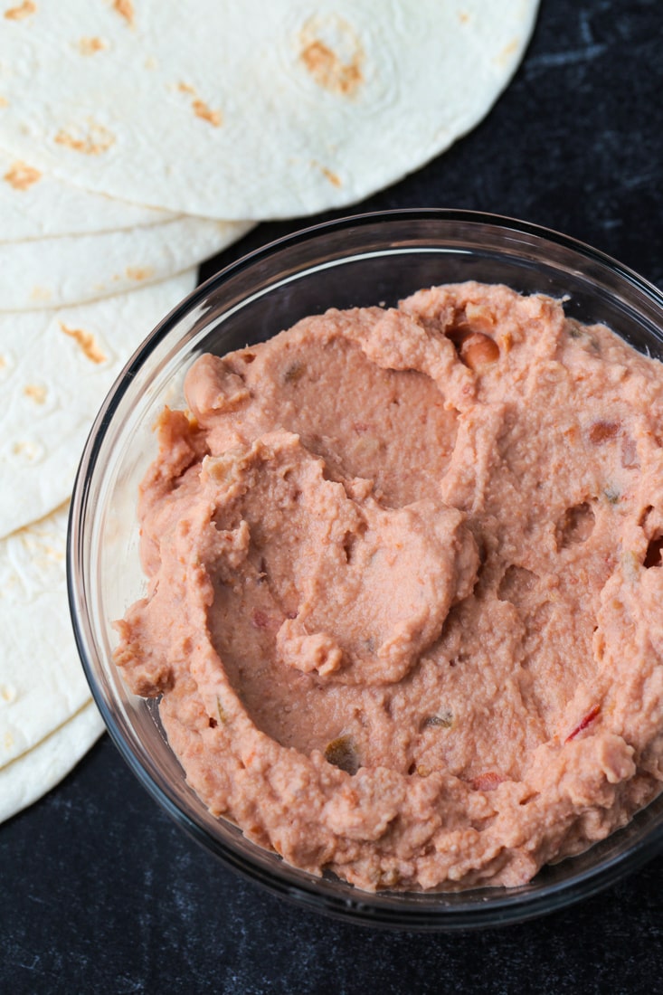 A glass bowl with season refried beans in it, with tortillas in the background