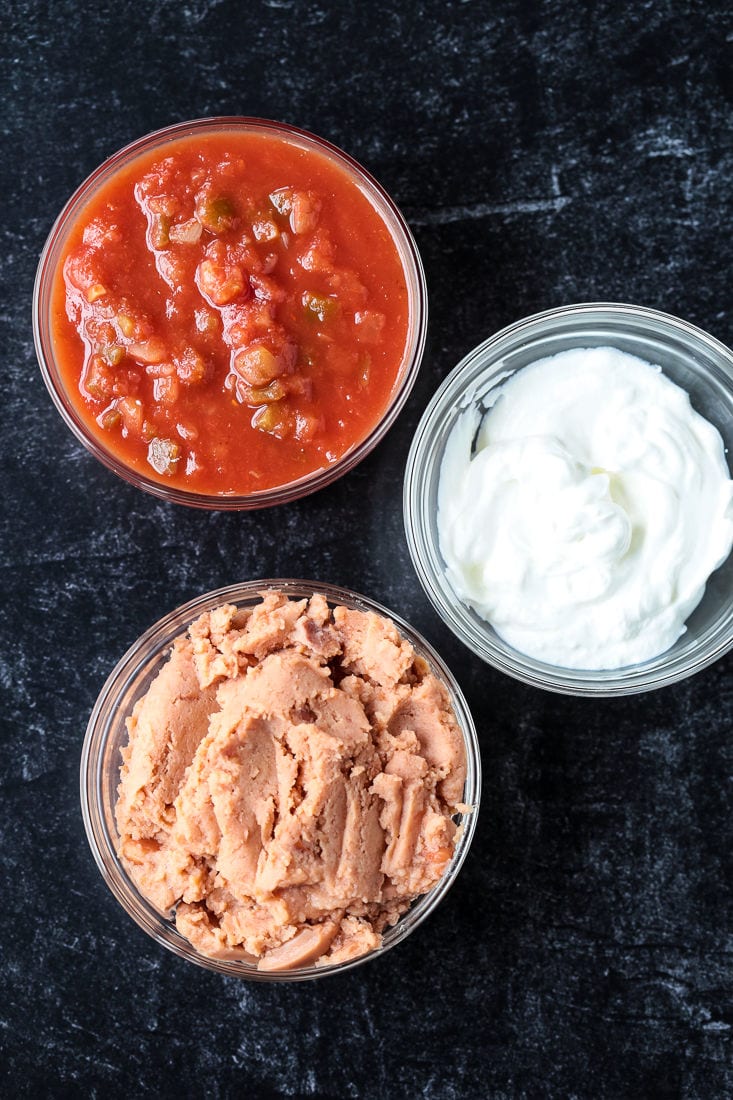 A bowl of refried beans, next to a bowl of sour cream, next to a bowl of salsa
