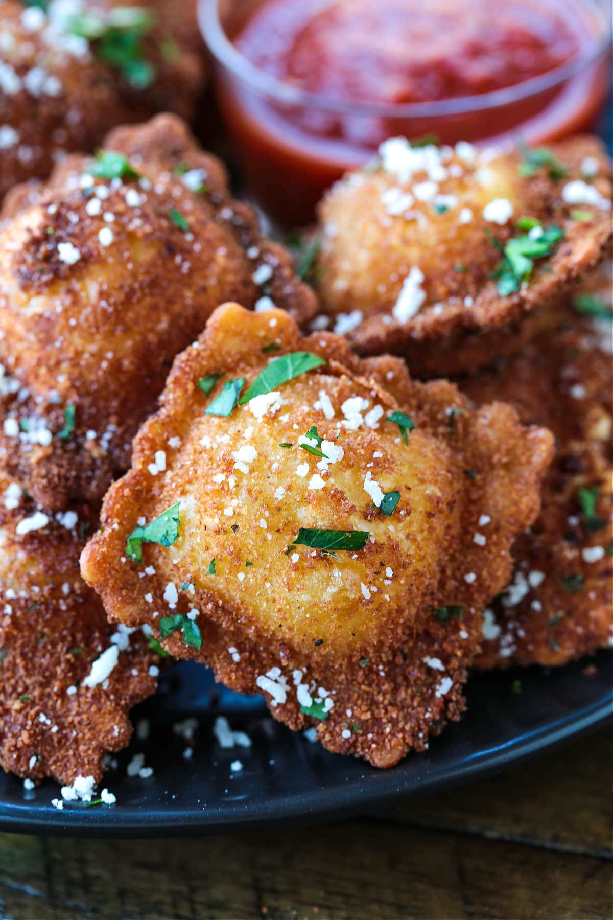 This fried ravioli recipe is crispy on the outside and soft and cheesy on the inside! Dip in marinara sauce for an addicting party appetizer! Close up of a fried ravioli with parsley and parmesan cheese on it, with more ravioli in the background