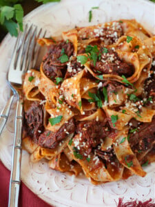 A plate of short rib ragu with pappardelle, topped with parsley and parmesan cheese, with a fork