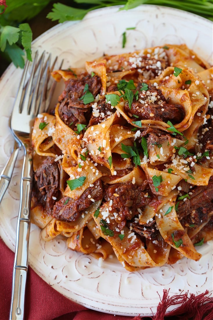 A plate of short rib ragu with pappardelle, topped with parsley and parmesan cheese, with a fork