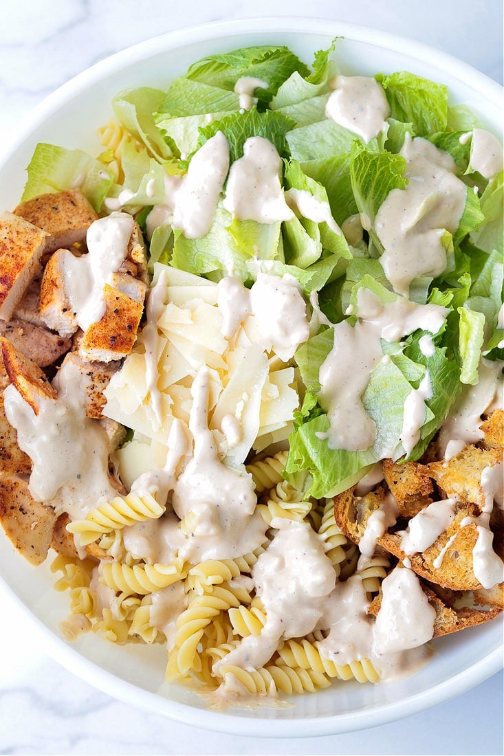 a salad bowl with lettuce, chicken, pasta and croutons, drizzled with dressing