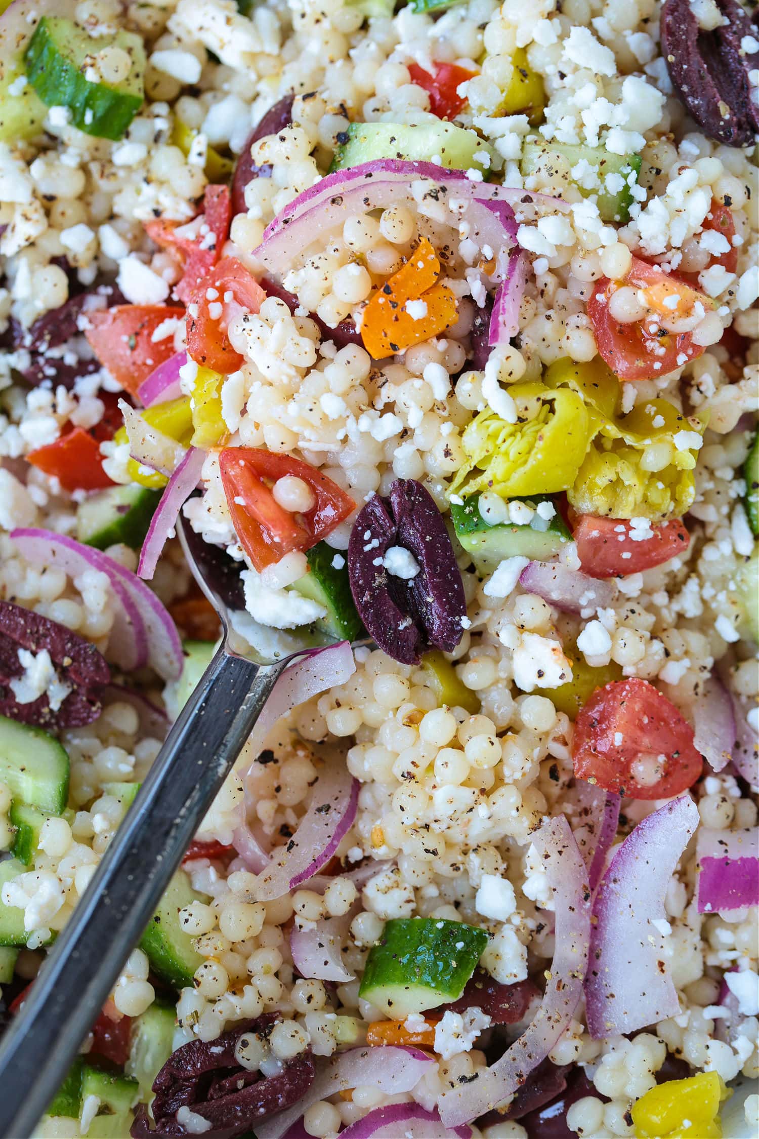 This Greek Couscous Salad is bursting with fresh flavors of ripe tomatoes, tangy olives, and creamy feta tossed with fluffy couscous. greek couscous salad in bowl with spoon