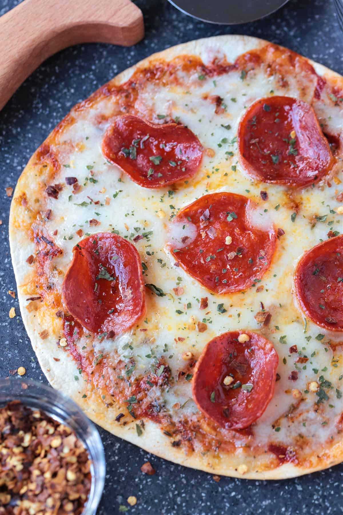 An Air Fryer Tortilla Pizza is perfect for an after school snack or an easy meal! A thin, crispy crust with gooey cheese ready in minutes! tortilla pizza with pepperoni on board with crushed red pepper