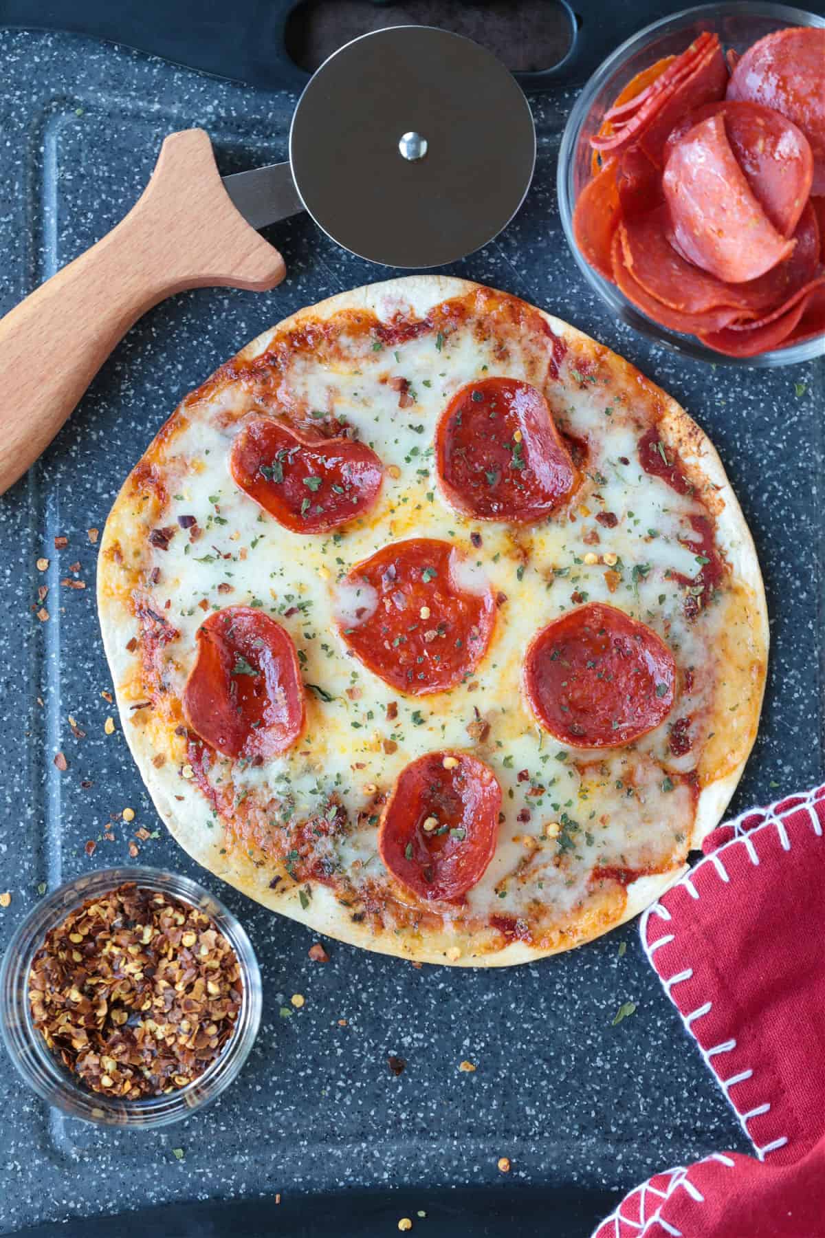An Air Fryer Tortilla Pizza is perfect for an after school snack or an easy meal! A thin, crispy crust with gooey cheese ready in minutes! tortilla pizza with pepperoni on board with pizza cutter