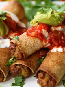 beef taquitos stacked on a plate with sour cream, salsa and guacamole