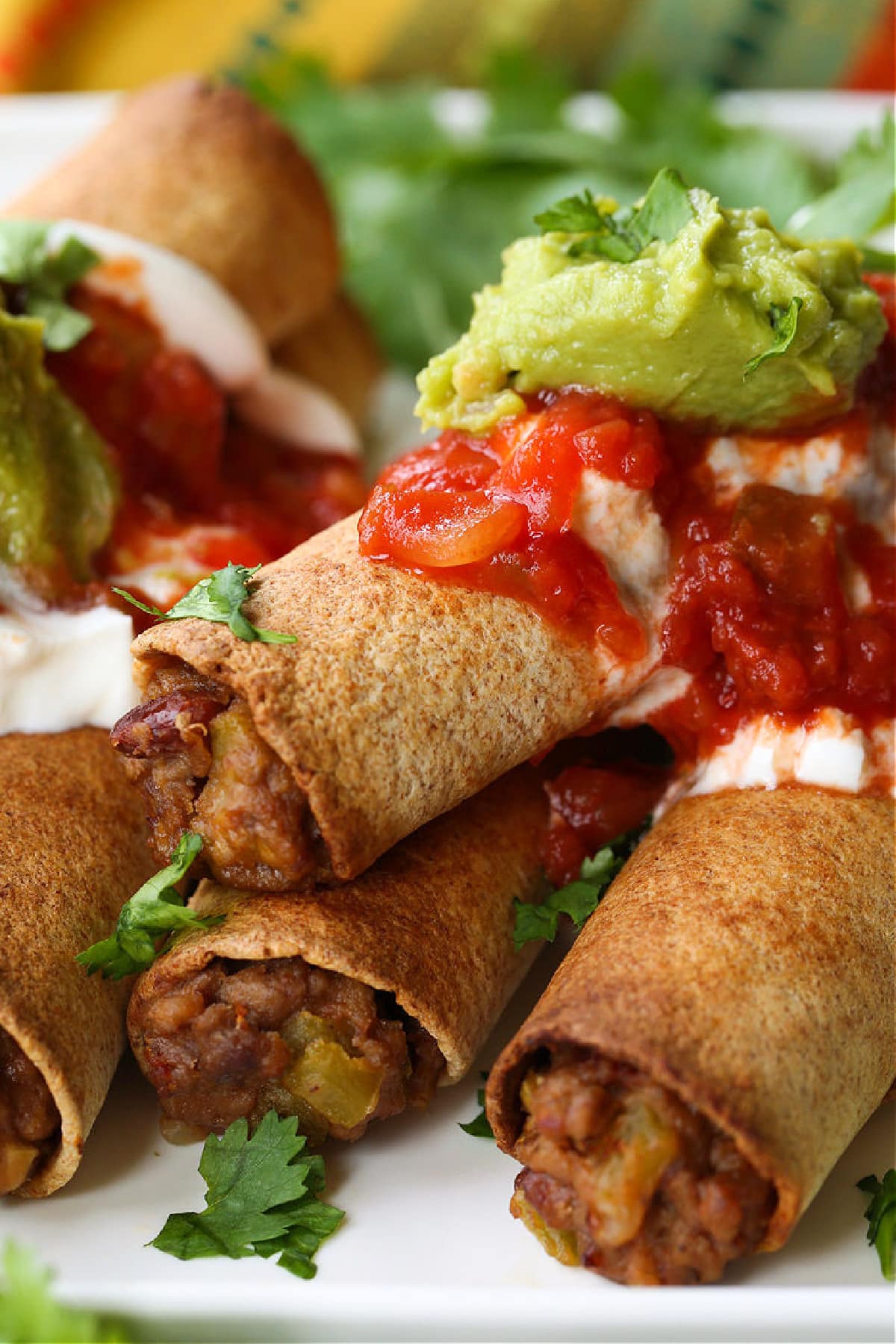 beef taquitos stacked on a plate with sour cream, salsa and guacamole