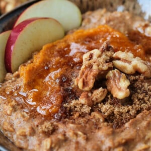 A bowlful of pumpkin oatmeal topped with apples, walnuts and pumpkin puree, with a spoon.