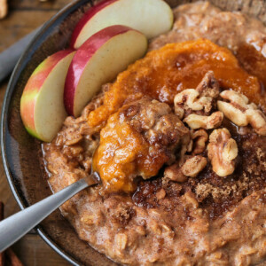 A bowlful of pumpkin oatmeal topped with apples, walnuts and pumpkin puree, with a spoon.