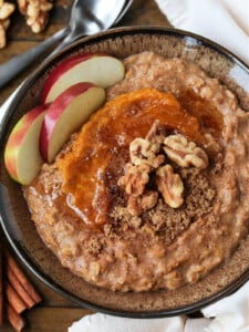 A bowlful of pumpkin oatmeal topped with apples, walnuts and pumpkin puree, with a spoon.