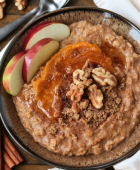 A bowlful of pumpkin oatmeal topped with apples, walnuts and pumpkin puree, with a spoon.