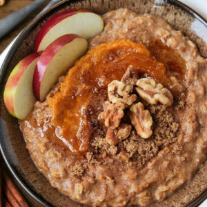A bowlful of pumpkin oatmeal topped with apples, walnuts and pumpkin puree, with a spoon.