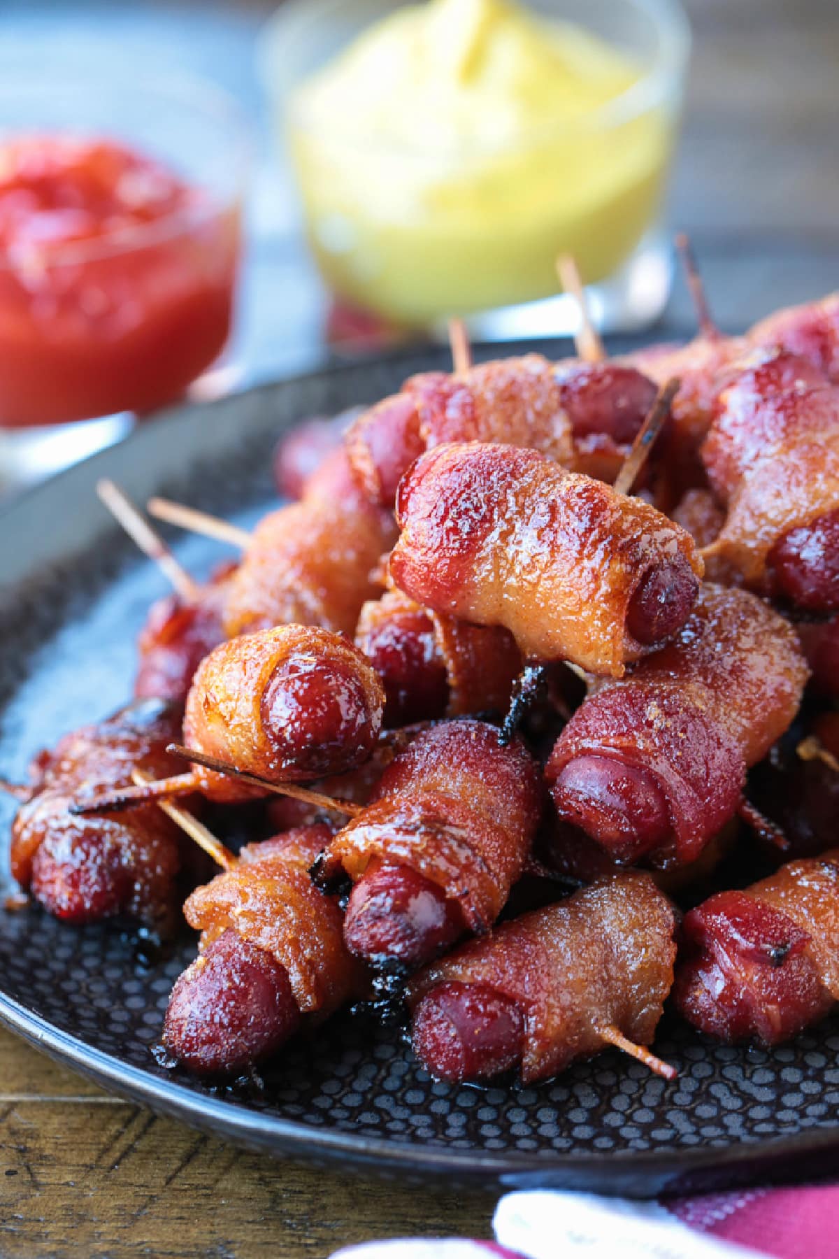 Bacon Wrapped Smokies are an addicting one-bite appetizer that everyone loves! Perfect for game days, parties and holidays! bacon wrapped mini hot dogs on plate with sauces