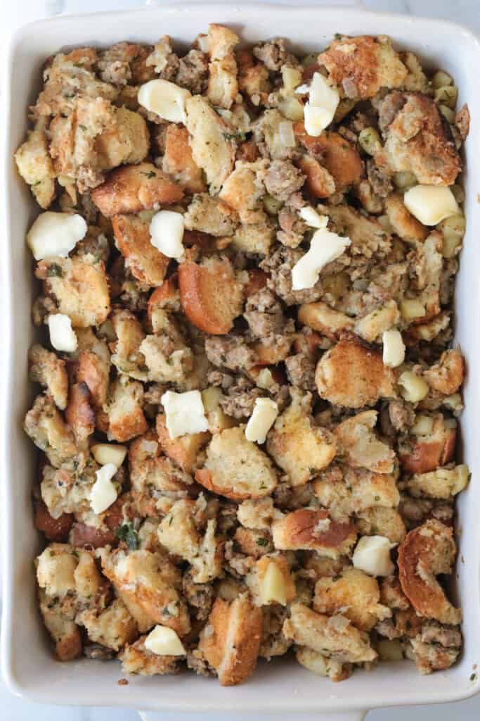 Our Sausage Stuffing Recipe is a favorite at Thanksgiving. Tons of sausage, sweet apples and buttery, toasted bread. We crave it all year! stuffing in a baking dish dotted with butter