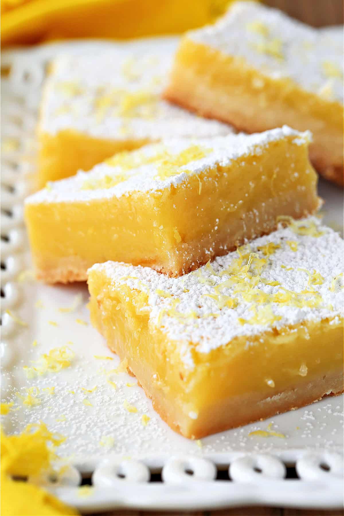 Our Limoncello Lemon Bars are a fun party dessert! Zesty lemon bars with a thick, buttery shortbread topped with a sweet and creamy custard. lemon bars stacked on white platter with powdered sugar