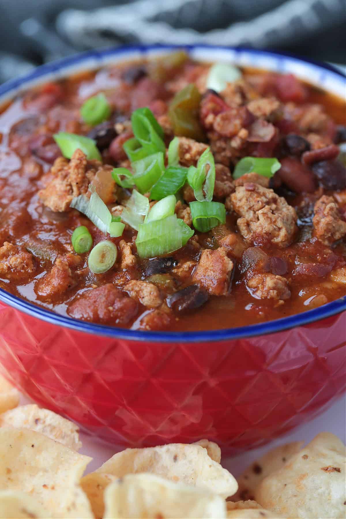 This slow cooker turkey chili is packed with lean ground turkey, vegetables and vibrant spices. Perfect for easy dinners or game days! turkey chili in a red bowl with scallions and chips on the side