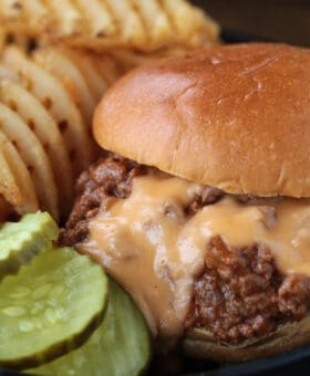 close up of cheesy sloppy joe on black plate with pickles and waffle fries