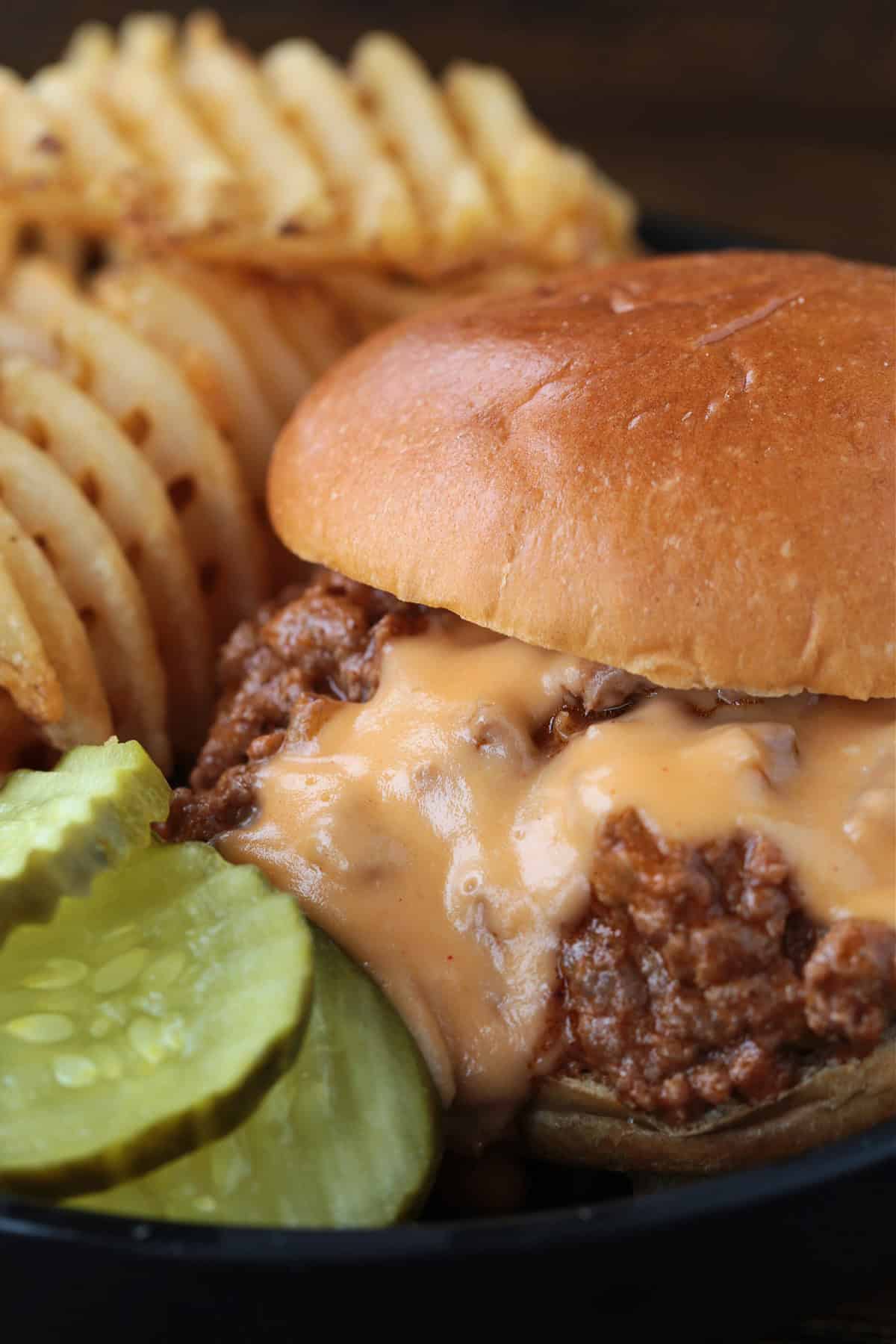 close up of cheesy sloppy joe on black plate with pickles and waffle fries