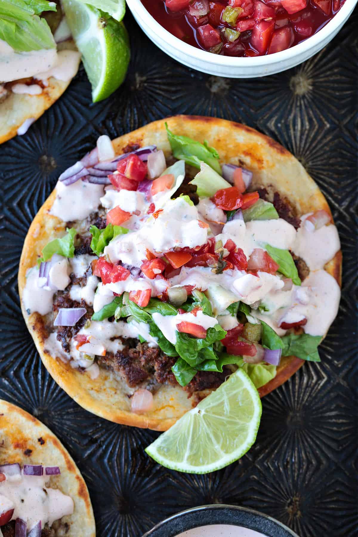 Smash Tacos are the key to fun dinners that everyone loves. Seasoned ground beef cooked on a crispy tortilla with your favorite toppings! smash taco with toppings and sauce and limes on the side