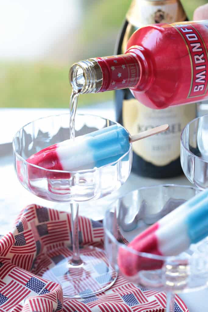This Bomb Pop Cocktail is the easiest summer drink you'll ever make! The popsicle melts into the champagne for added fun and flavor! berry vodka pouring into champagne glass with ice pop
